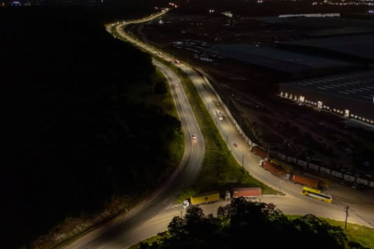 Vista aérea noturna da Avenida BYD em Camaçari com iluminação em LED, veículos em circulação e área industrial ao lado da via.