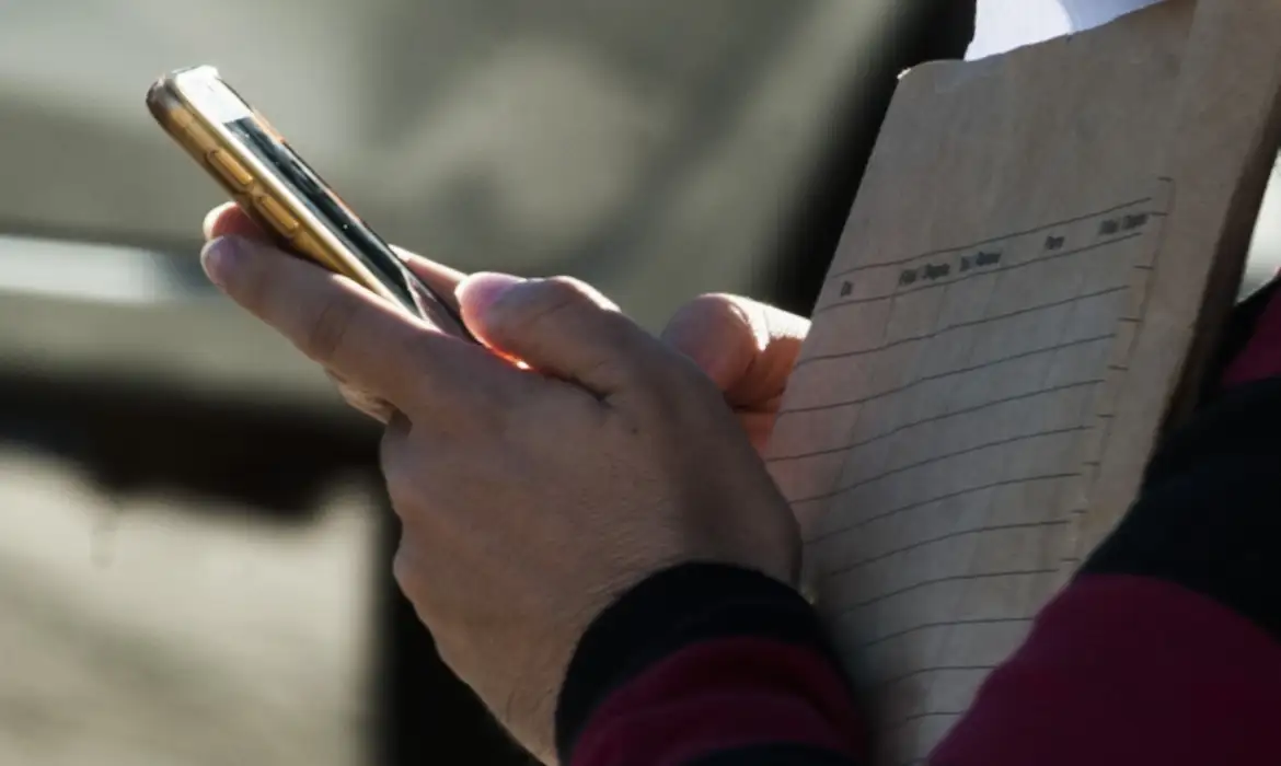 Pessoa segura celular ao lado de prancheta com papel, em imagem relacionada ao BB Governo Digital em Ipatinga