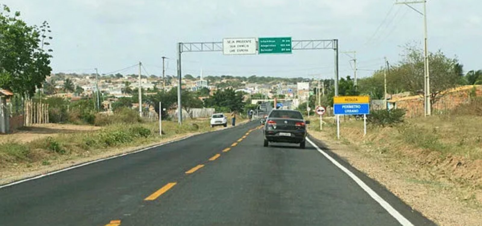 Trecho de rodovia na Bahia com pista asfaltada e acesso urbano em imagem de obra viária.