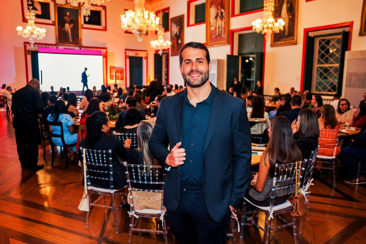 Homem posa em primeiro plano durante evento institucional realizado em salão histórico com público ao fundo.