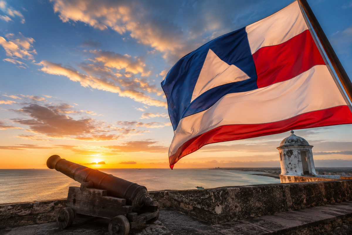 Bandeira da Bahia hasteada ao lado de um canhão histórico durante o pôr do sol, em imagem que representa o 2 de Julho, data da Independência da Bahia.